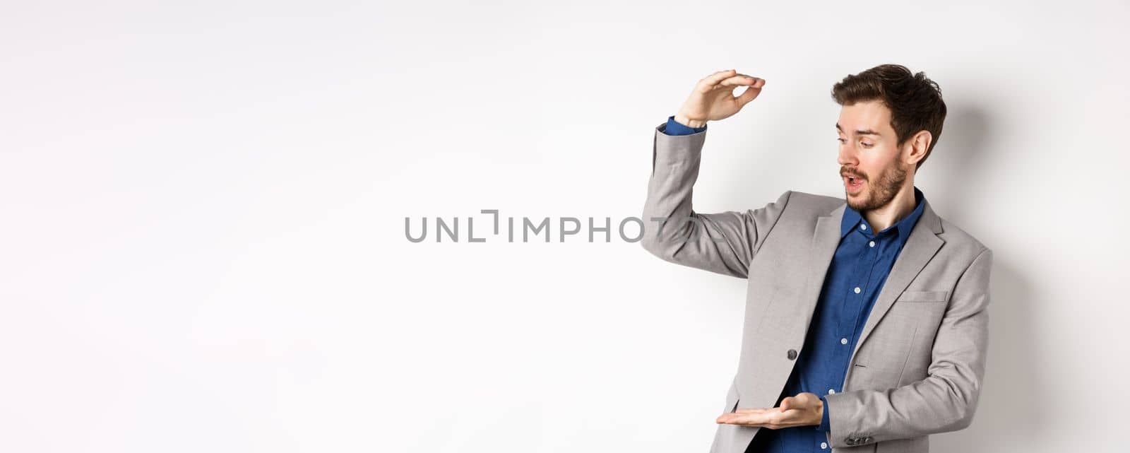 Businessman in suit showing large size and looking excited, making big money, standing on white background.