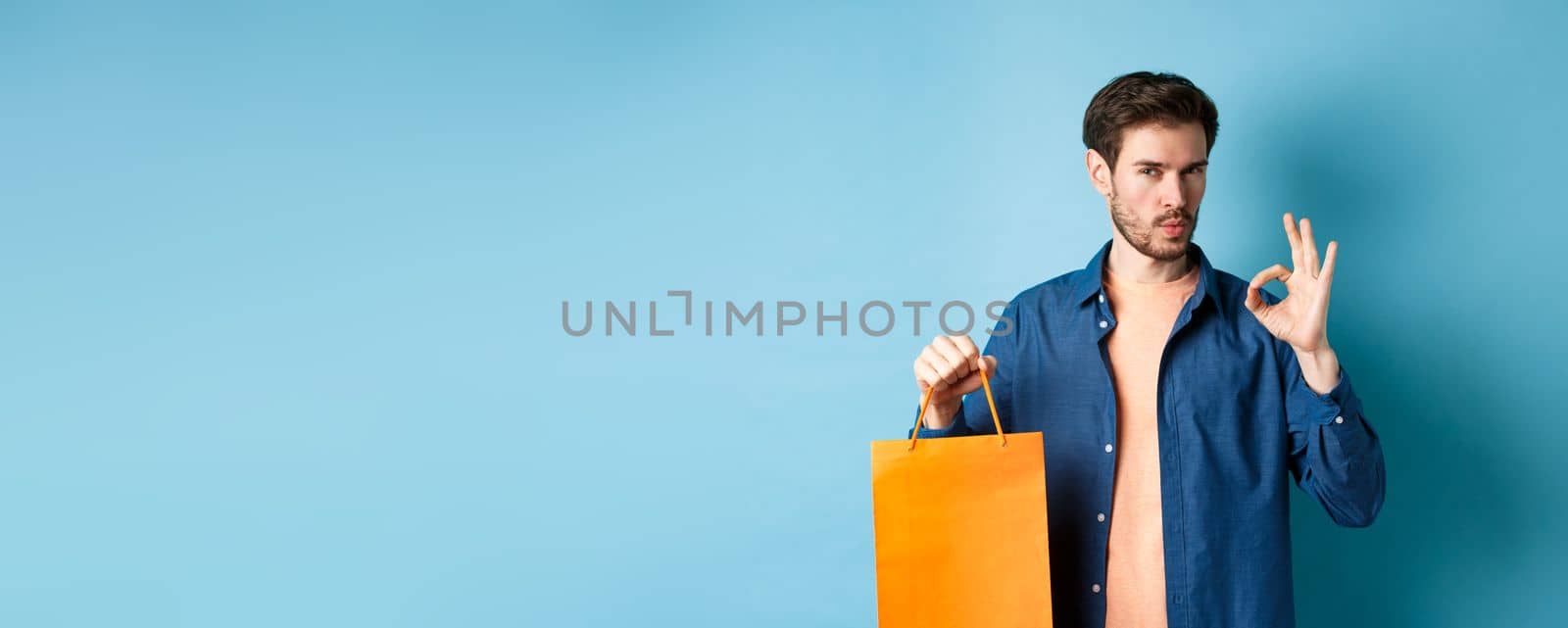 Handsome young man recommending store, showing orange shopping bag and OK sign, standing on blue background.