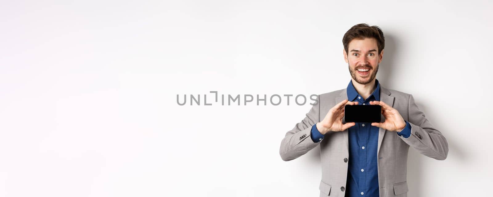 E-commerce and online shopping concept. Excited young businessman in suit showing empty smartphone, screen demonstrate app achievement, white background by Benzoix