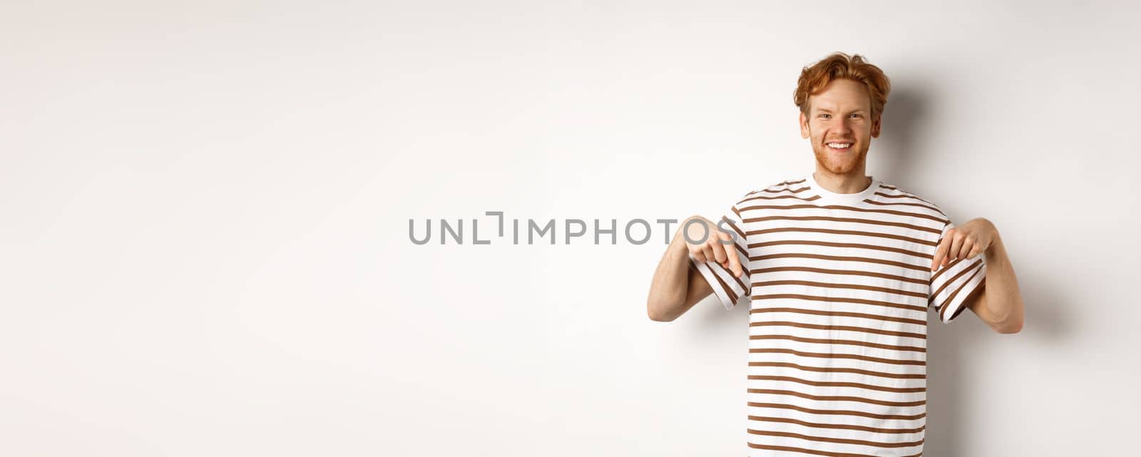 Smiling young man with messy red hair and beard, pointing fingers down, recommending shop, standing over white background by Benzoix