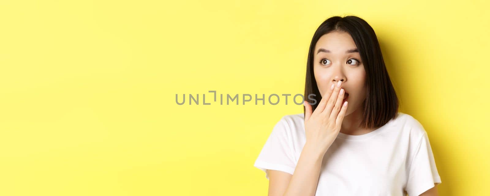 Close up of pretty asian girl looking shocked, gasping and covering mouth with hand, stare left at promotion, standing over yellow background.