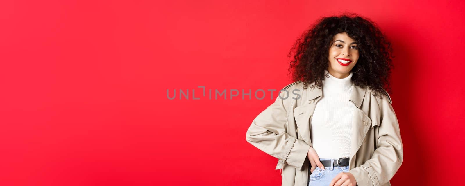 Modern woman with curly hair, wearing outdoor clothes, going for walk, standing against red background by Benzoix