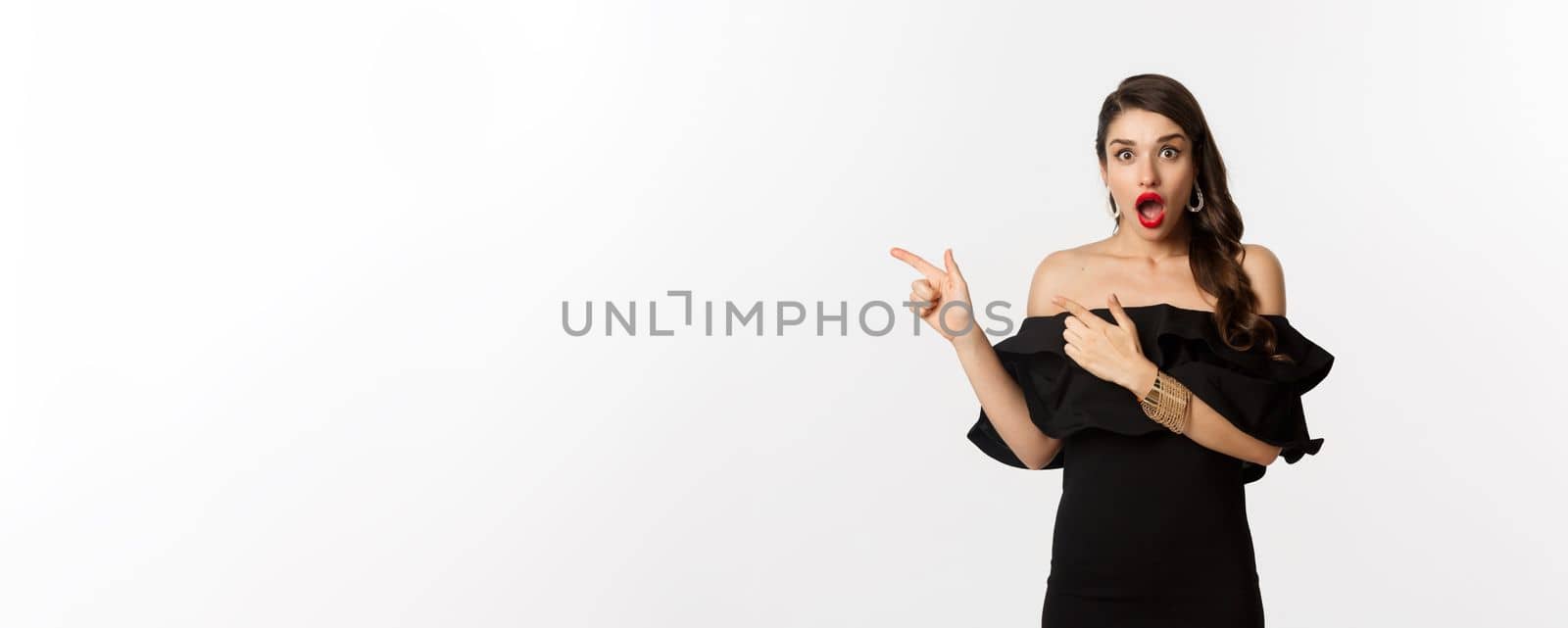 Fashion and beauty. Surprised woman in black glamour dress pointing fingers left, showing advertisement and staring amazed, white background.