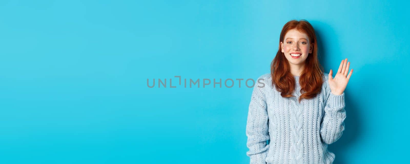 Friendly redhead teenage girl saying hi, waving hand in hello gesture and smiling, standing against blue background.