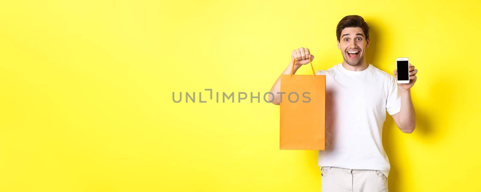 Surprised man holding shopping bag and showing smartphone screen, concept of mobile banking and app achievements, yellow background by Benzoix