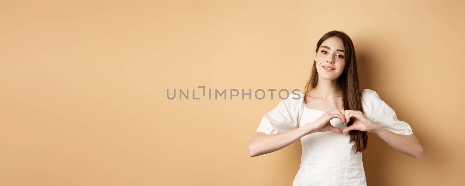 Beautiful caucasian woman say I love you, showing heart gesture and smile at camera, beige background. Concept of romance and Valentines day.