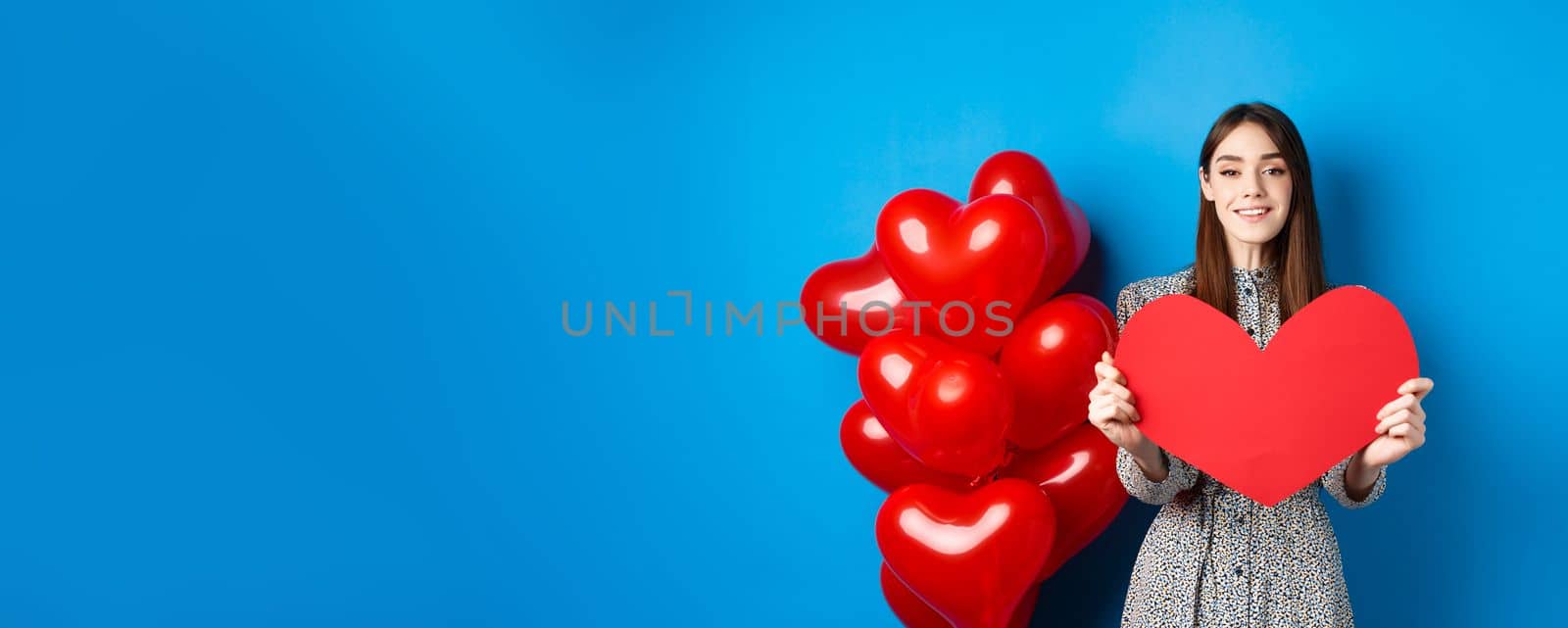 Valentines day. Lovely young woman in dress celebrating lovers holiday, showing valentine card and smiling, standing near red heart balloons on blue background by Benzoix