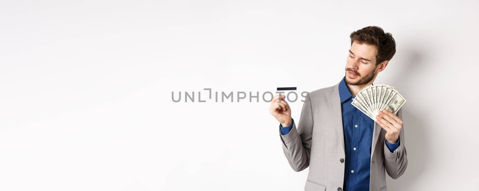 Successful man holding dollar bills and looking at plastic credit card, pleased with bank, standing on white background in business suit.