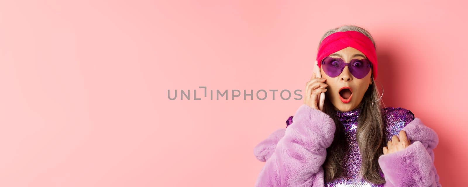 Fashion. Close-up of stylish asian old woman hear shocking news on smartphone, gasping and staring amazed at camera, wearing trendy sunglasses, pink background by Benzoix
