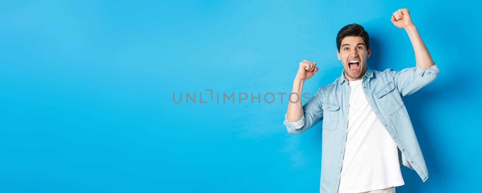 Cheerful guy making fist pumps and rooting for someone, shouting for joy, triumphing over win, standing against blue background.