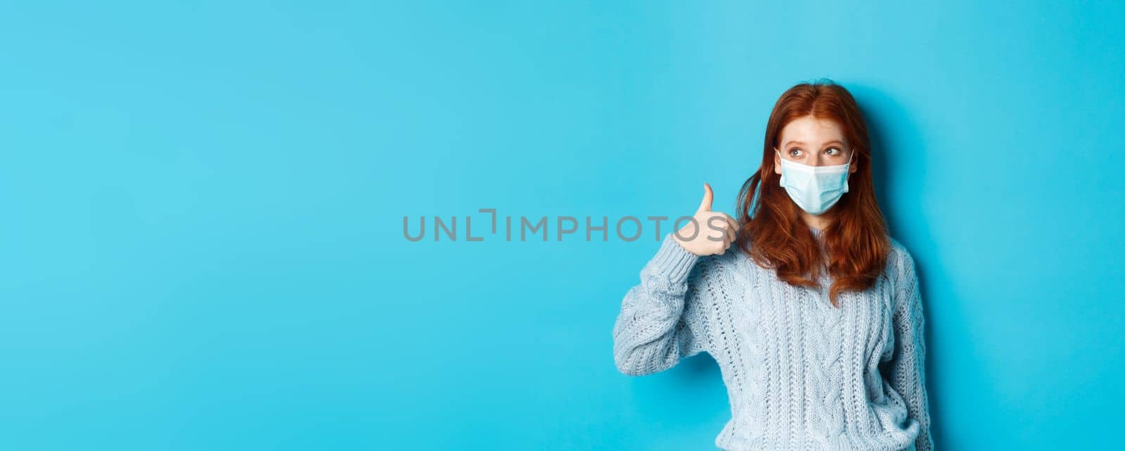 Winter, covid-19 and social distancing concept. Satisfied young redhead woman in face mask showing alright, thumb up gesture and looking left at promo, blue background by Benzoix