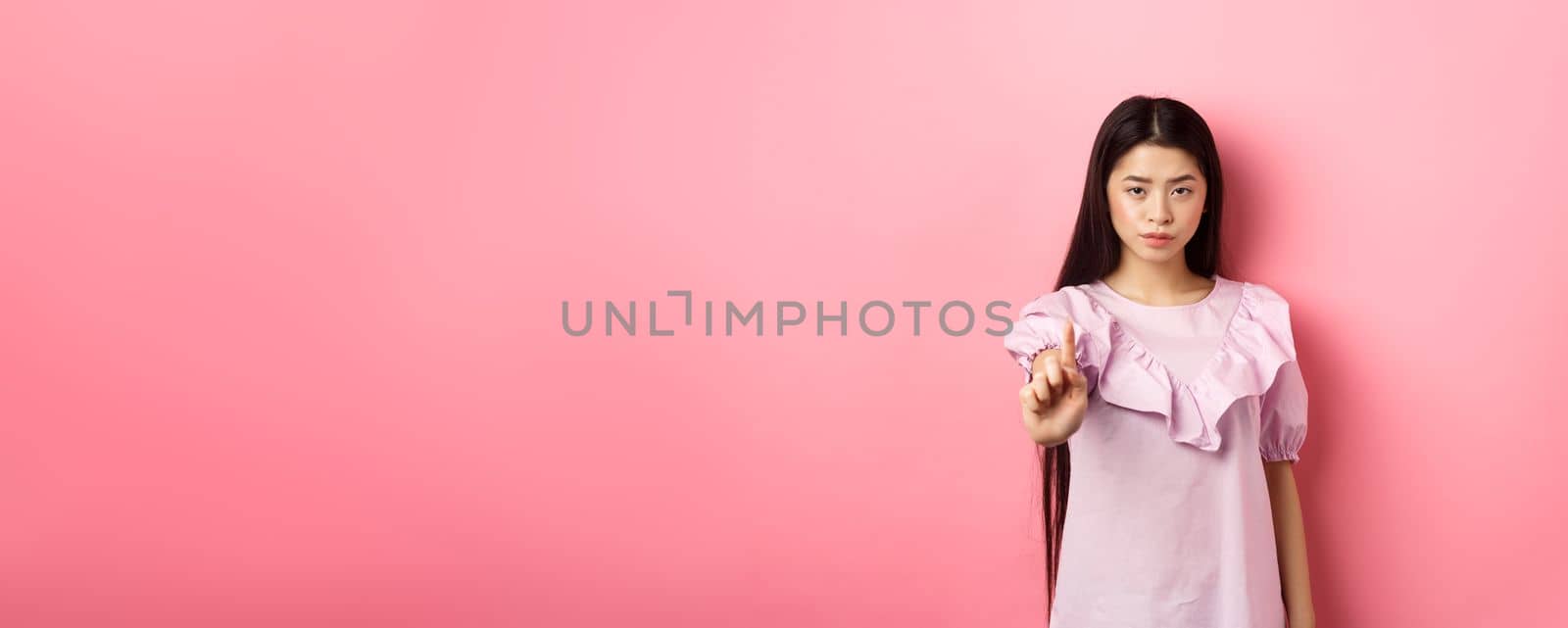 She says no. Serious asian girl shaking finger in stop gesture, prohibit and disagree with person, scolding bad behaviour, standing against pink background.