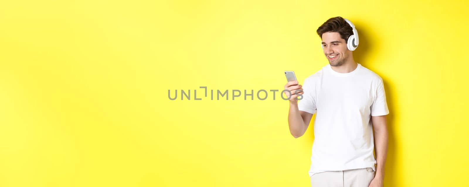 Handsome modern guy choosing playlist on smartphone, wearing headphones, standing over yellow background by Benzoix