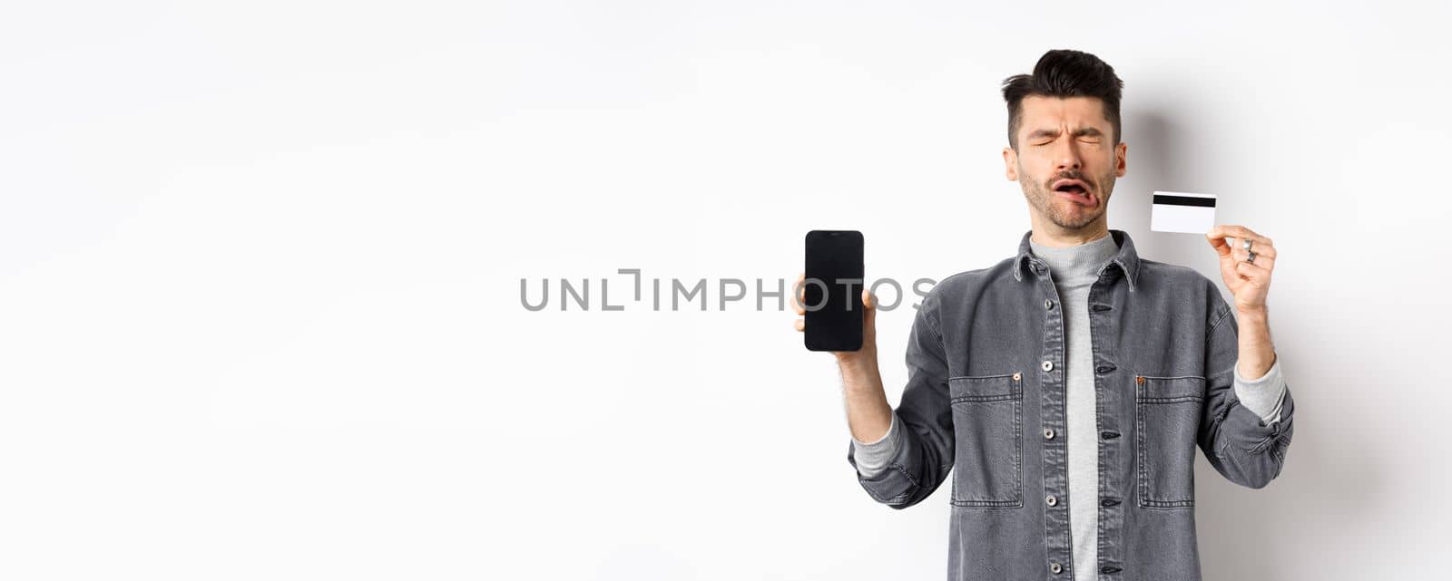Sad crying man show empty mobile phone screen and plastic credit card, standing miserable on white background.