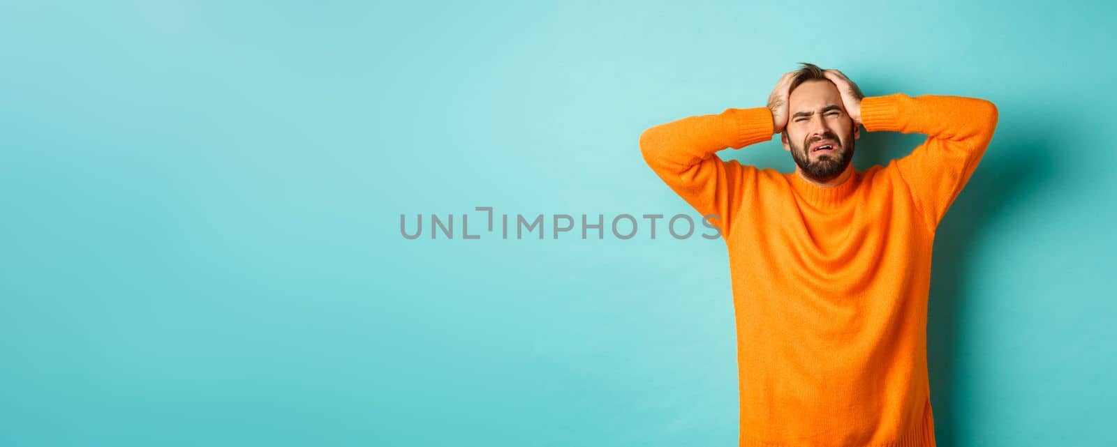 Image of frustrated man in orange sweater, holding hands on head and feeling distressed, standing anxious against turquoise background by Benzoix