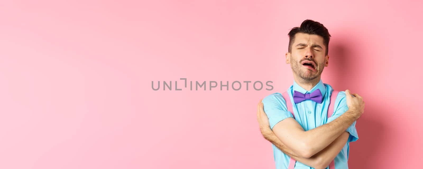 Crying guy hugging himself, feeling sad and lonely, standing unhappy on pink background. Copy space