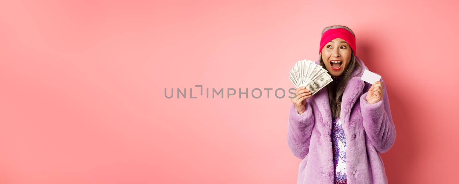Shopping and fashion concept. Asian senior woman scream happy like winner, holding dollars money and plastic credit card, looking excited, pink background by Benzoix