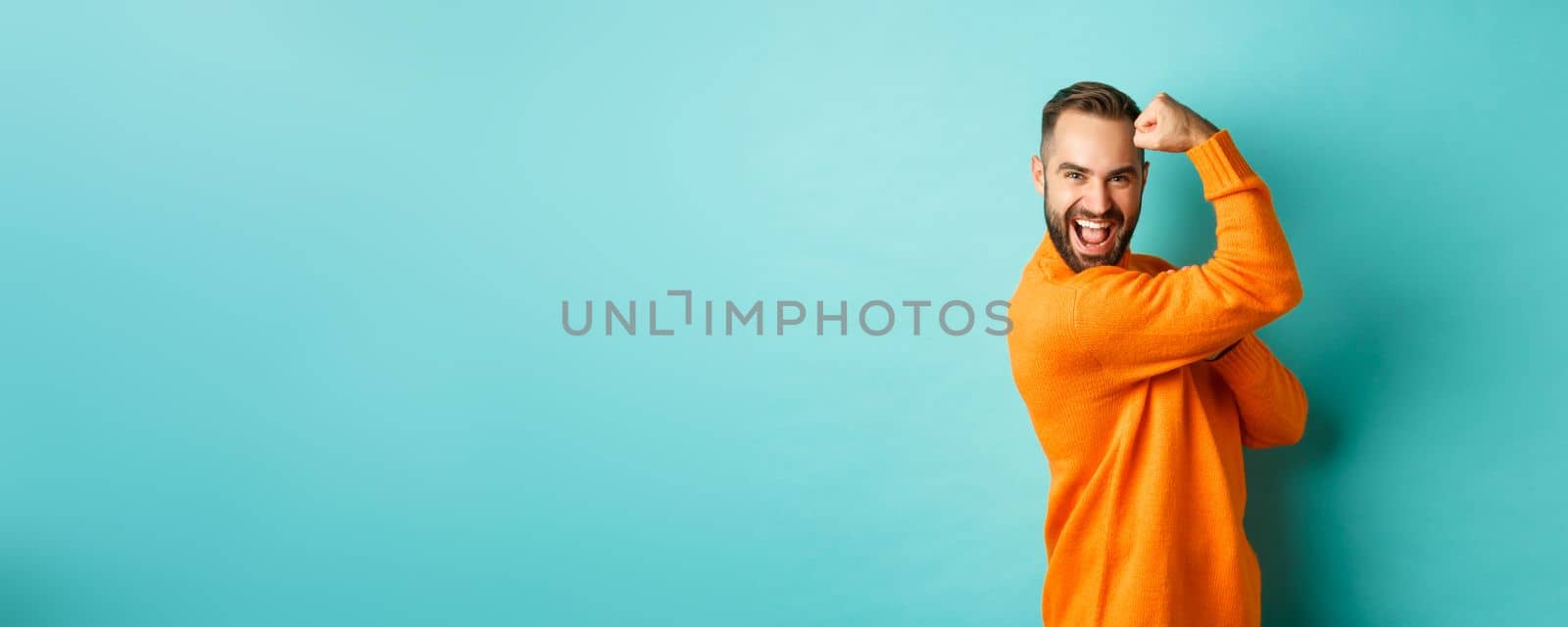 Confident handsome man show-off his muscles, flex biceps, feeling strong, standing over light blue background.