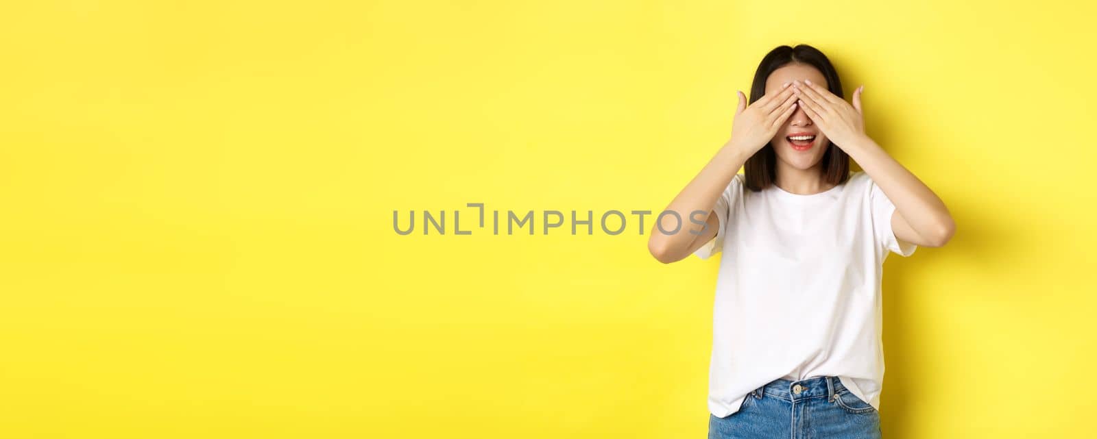 Happy asian woman waiting for surprise gift, close eyes and smiling, expecting something, standing over yellow background.