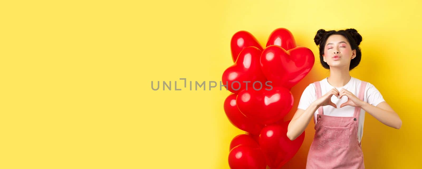 Romantic girl close eyes and pucker lips for kiss, showing I love you heart gesture, standing near cute red balloons, kissing lover over yellow background.