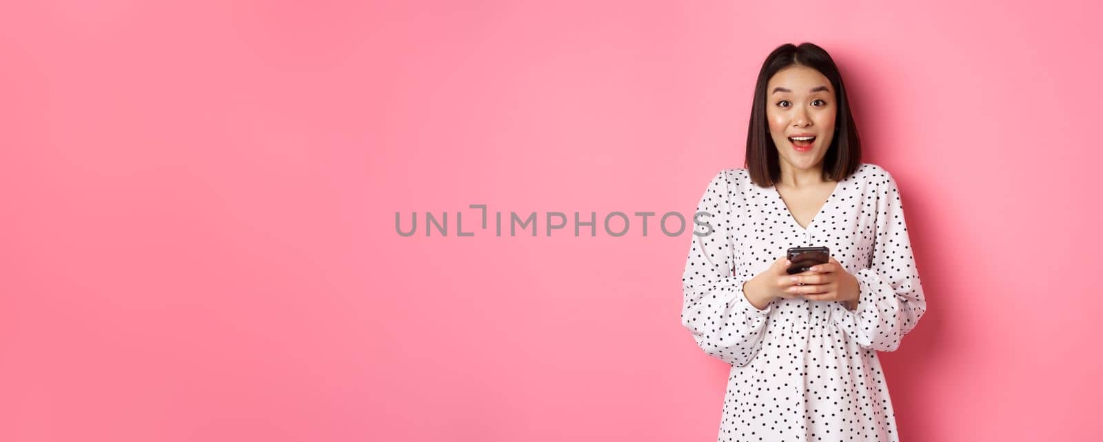 Online shopping. Amazed asian woman looking at camera with happy smile, making purchase with smartphone, using mobile phone app, standing over pink background by Benzoix