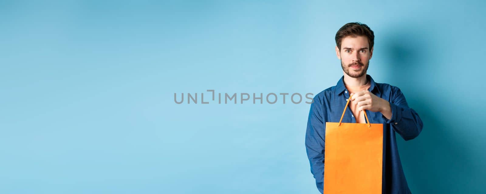 Handsome bearded man stretch out hand with orange shopping bag, making a gift, standing on blue background by Benzoix