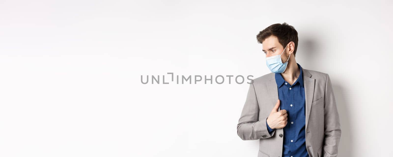 Covid, pandemic and business concept. Handsome male entrepreneur wearing stylish suit and medical mask, looking aside, white background.