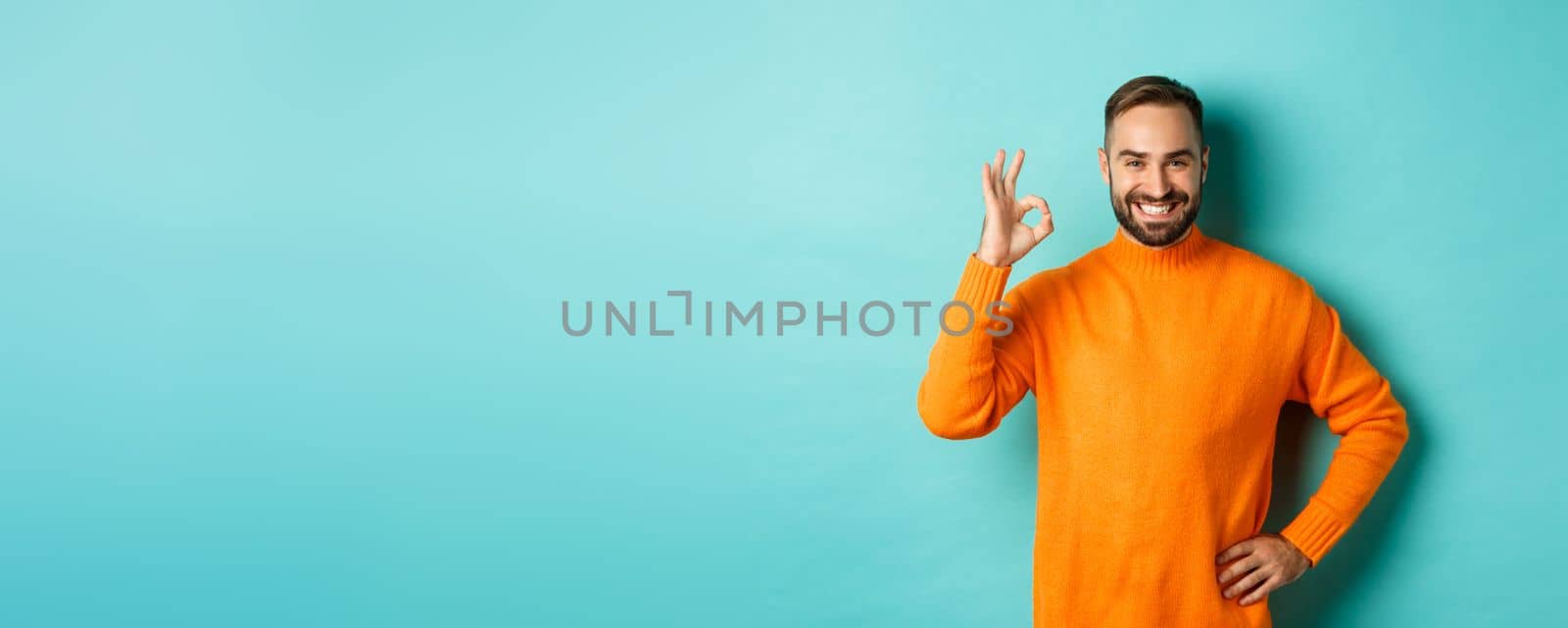 Image of confident handsome man showing ok sign, approve and agree, like something good, standing in orange sweater.