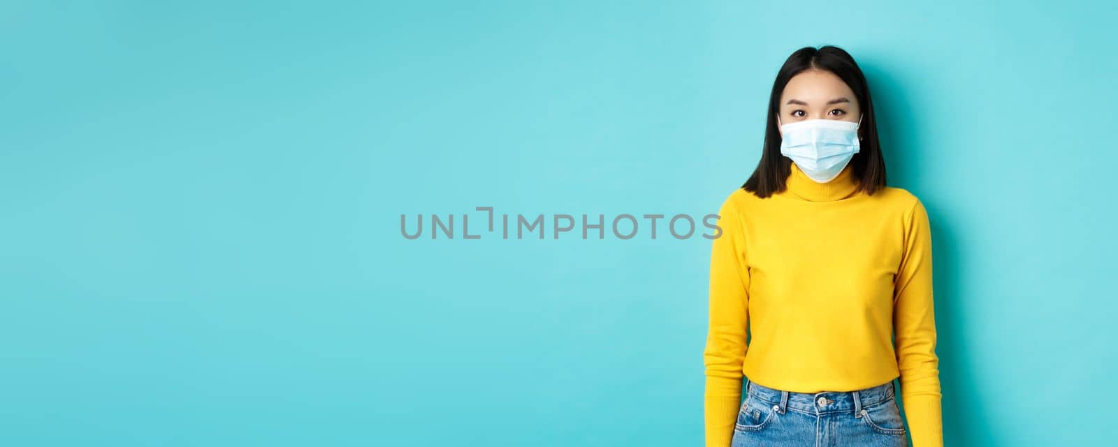Covid-19, social distancing and pandemic concept. Teenage asian woman in medical mask, looking at camera and standing against blue background in yellow sweater.