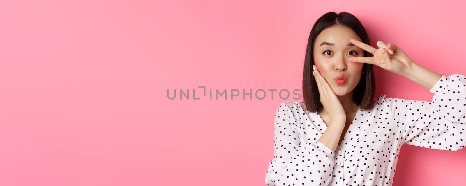 Beauty and lifestyle concept. Close-up of pretty asian woman showing peace sign and touching cheek, smiling happy at camera, standing over pink background.