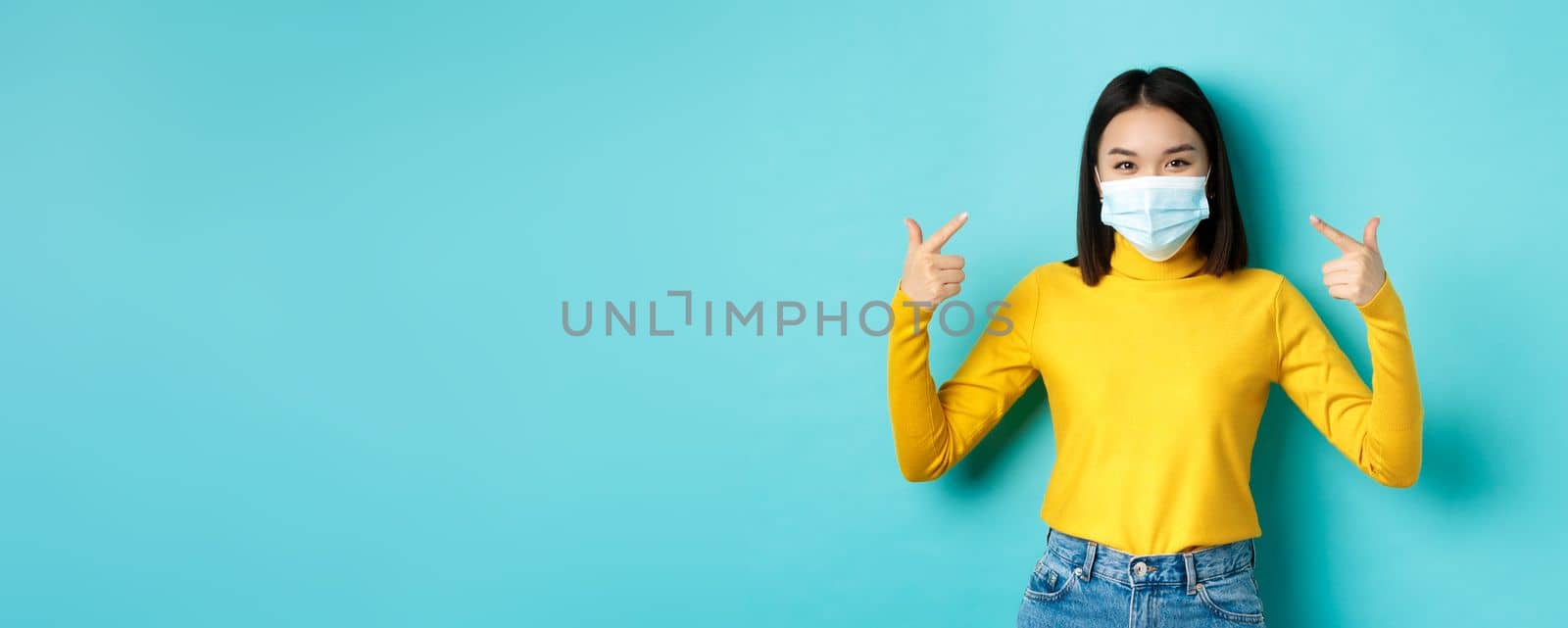 Covid-19, social distancing and pandemic concept. Young asian woman protect herself from coronavirus, pointing finger at her medical mask, standing over blue background.