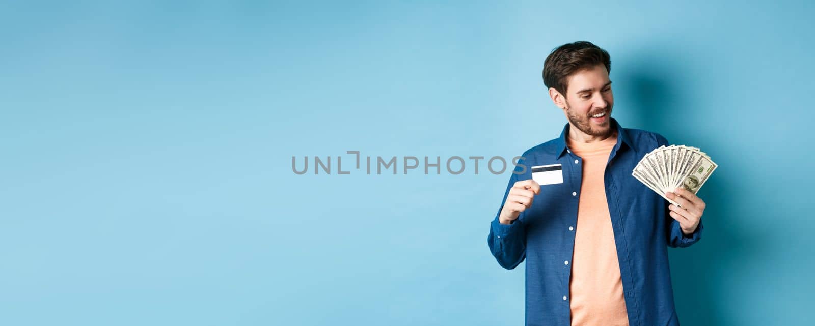 Smiling modern guy looking at cash and showing plastic credit card, standing on blue background. Copy space