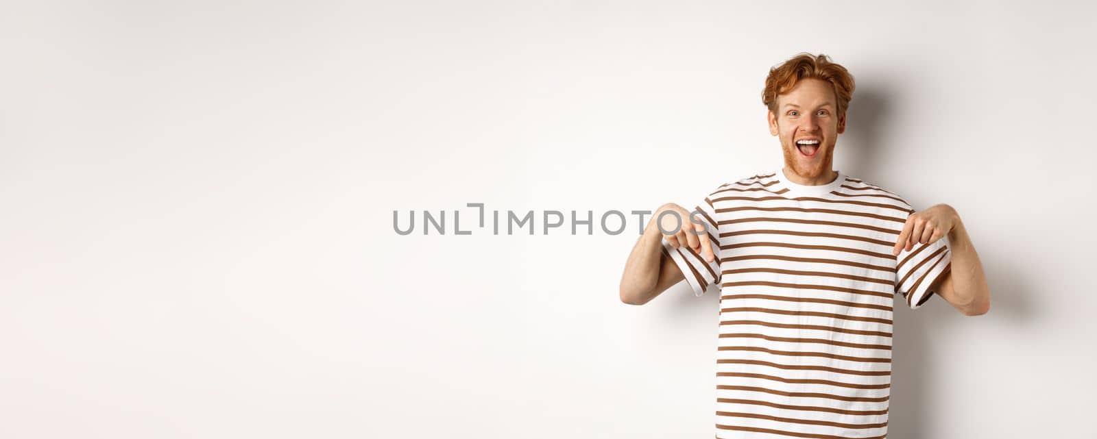 Enthusiastic young man with ginger hair and bristle showing advertisement, pointing fingers down and smiling, white background by Benzoix