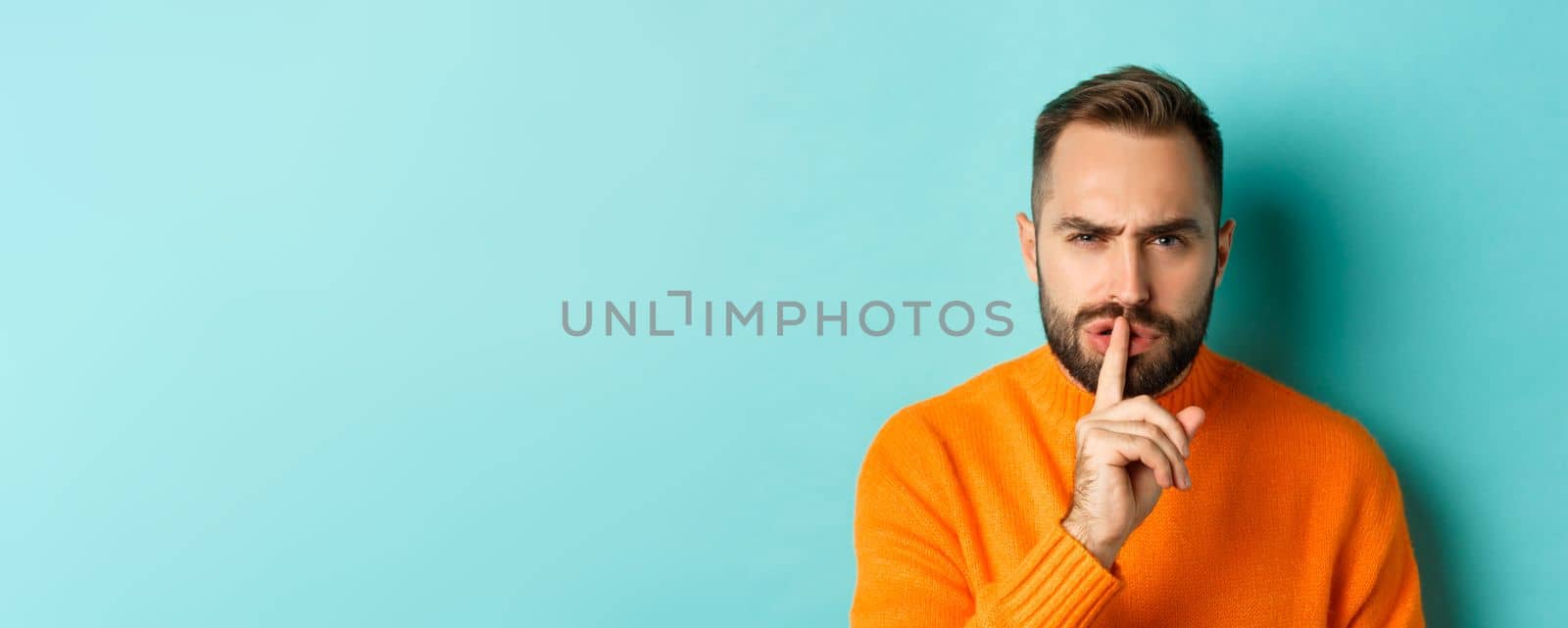 Close-up of annoyed frowning man shushing at camera, press finger to lips, showing taboo gesture, standing over light blue background by Benzoix