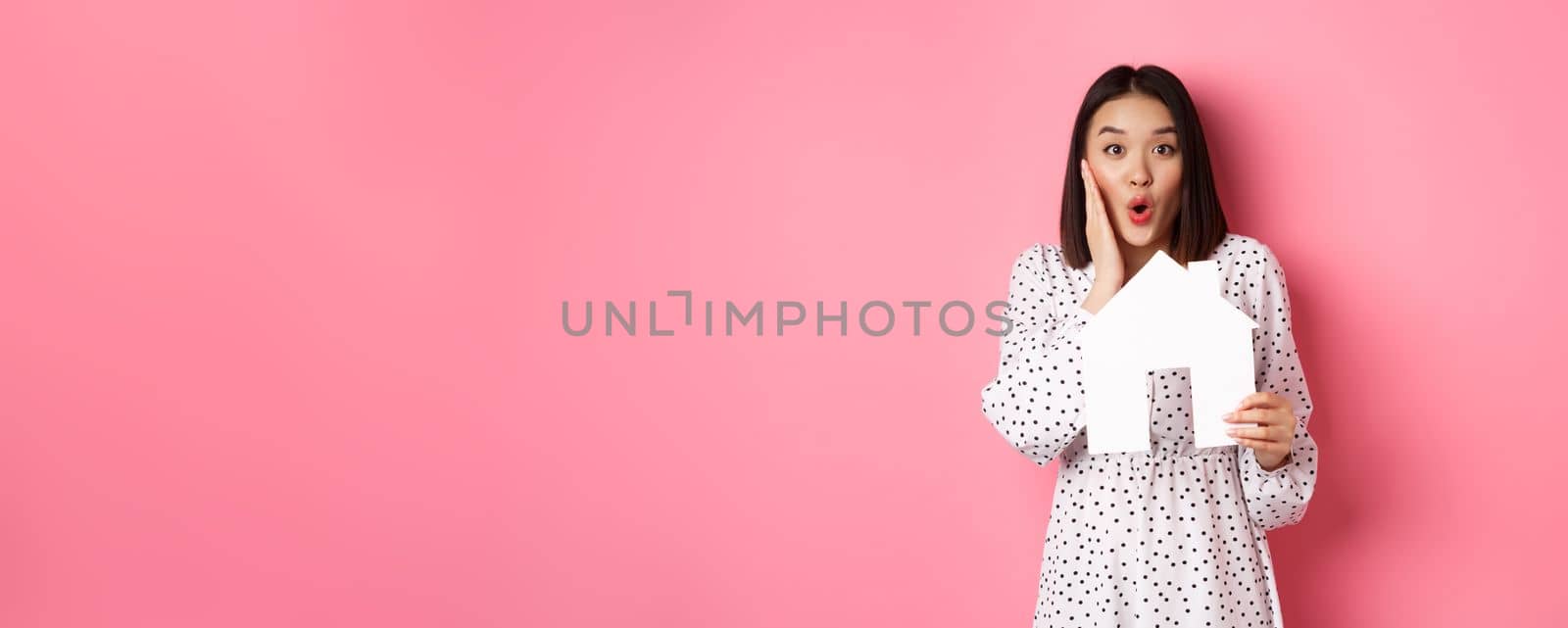 Real estate. Surprised and amazed asian woman staring at camera, saying wow and showing paper house model, standing over pink background by Benzoix