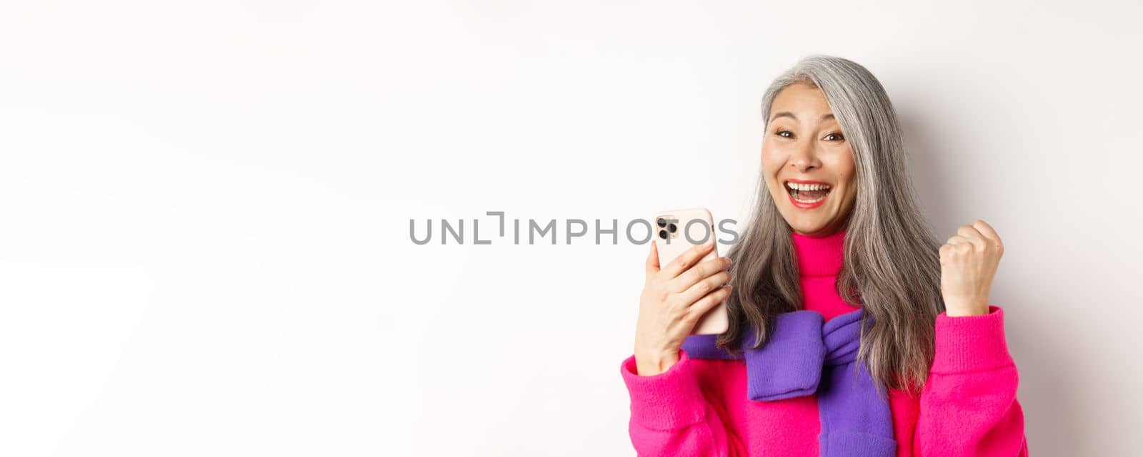 Online shopping. Close-up of cheerful asian elderly woman winning prize in internet, holding smartphone and making fist pump, standing over white background.