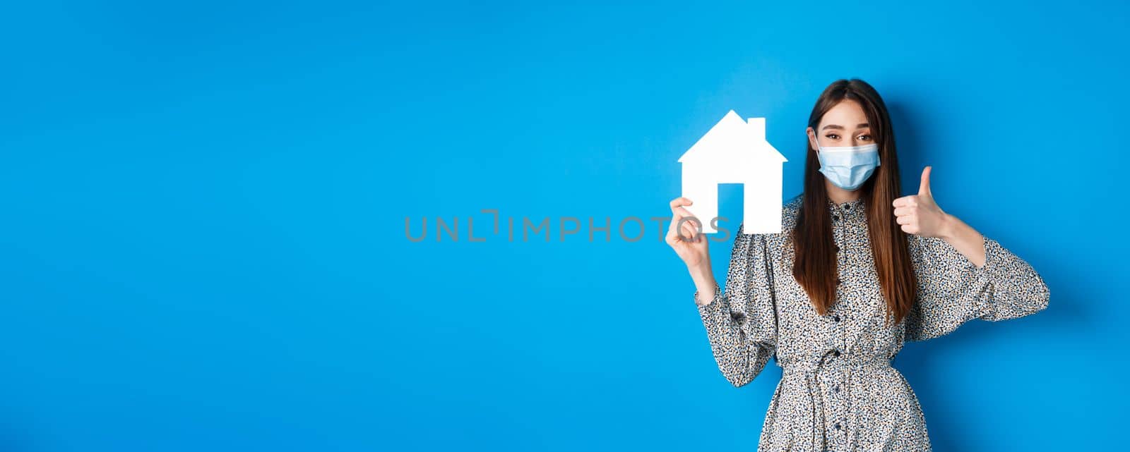 Real estate, covid-19 and pandemic concept. Cheerful woman in medical mask showing paper house cutout and thumbs up, recommend agency or realtor website, blue background.