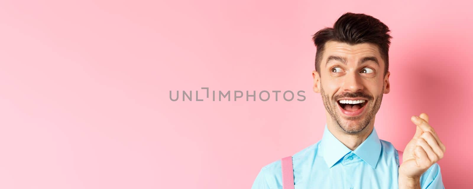 Valentines day concept. Romantic guy showing finger heart and looking left, smiling happy, standing on pink background dreamy.