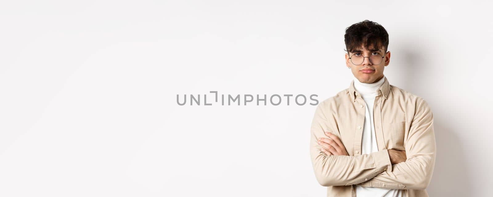 Image of stylish hipster guy in glasses feeling bored, looking unamused with pokerface, cross arms on chest and stand reluctant against white background.