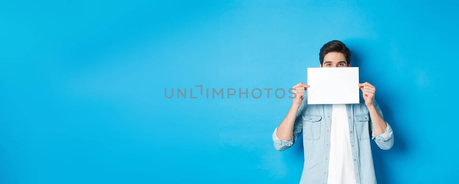 Sneeky handsome guy hiding face behind blank piece of paper for your logo, making announcement or showing promo offer, standing over blue background by Benzoix