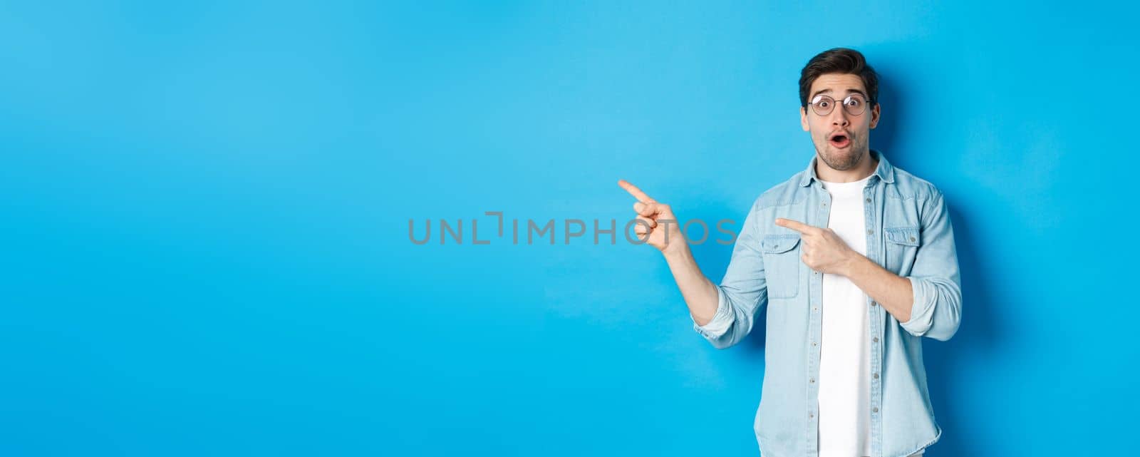 Surprised man in glasses pointing fingers left at copy space, asking question about product, looking intrigued, standing over blue background by Benzoix