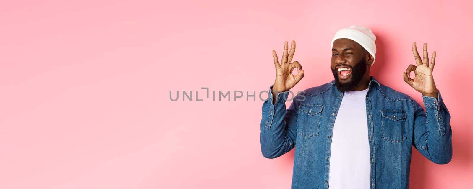 Satisfied african-american man in hipster beanie, showing okay signs in approval, praising good offer and saying yes, smiling pleased, standing over pink background by Benzoix