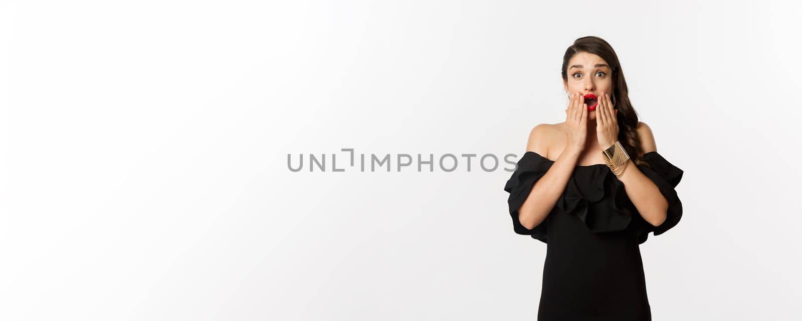 Fashion and beauty. Image of attractive female model in black dress reacting to announcement, looking amazed at camera, standing surprised over white background.