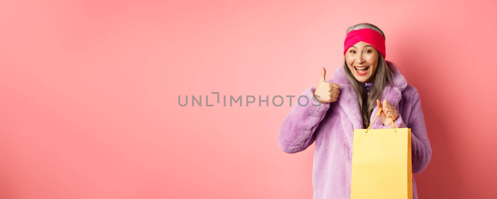 Shopping and fashion. Happy asian senior woman in stylish clothes holding paper bag from store, showing thumb-up in approval, pink background by Benzoix