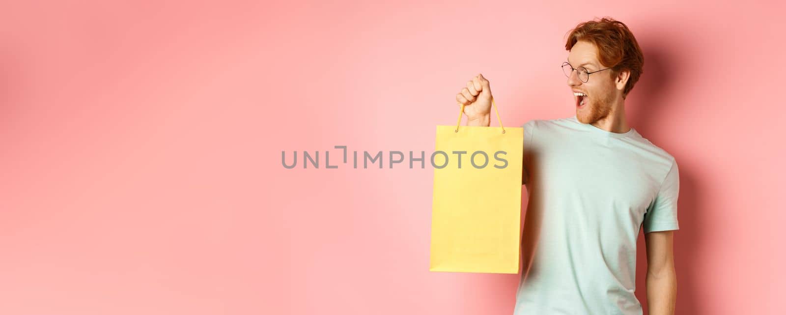 Handsome young man buying presents, holding shopping bag and looking amused, standing over pink background by Benzoix