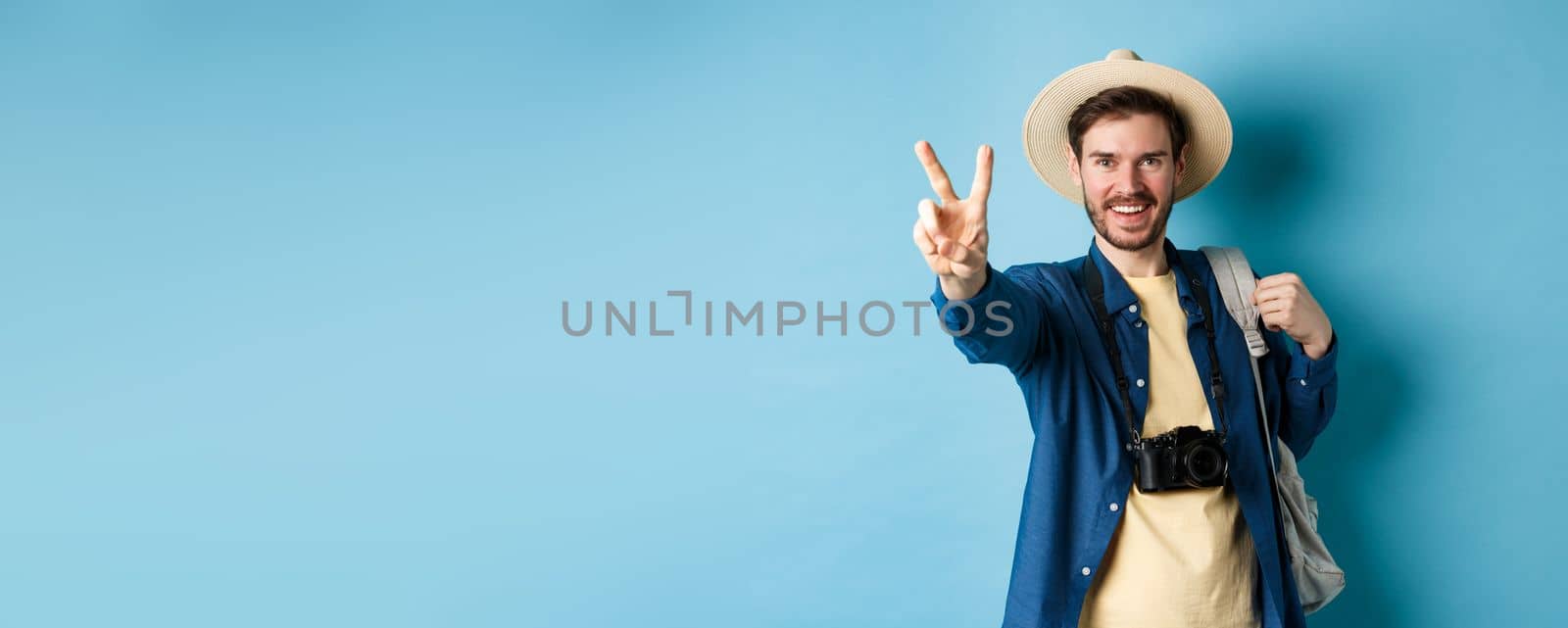 Handsome positive guy tourist showing peace sign on summer vacation, enjoying tropical holidays under sun, wearing straw hat, holding backpack and camera, blue background.