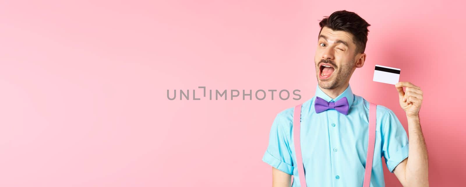 Shopping concept. Funny guy with moustache winking at camera, showing plastic credit card, recommending bank promo offer, standing on pink background by Benzoix