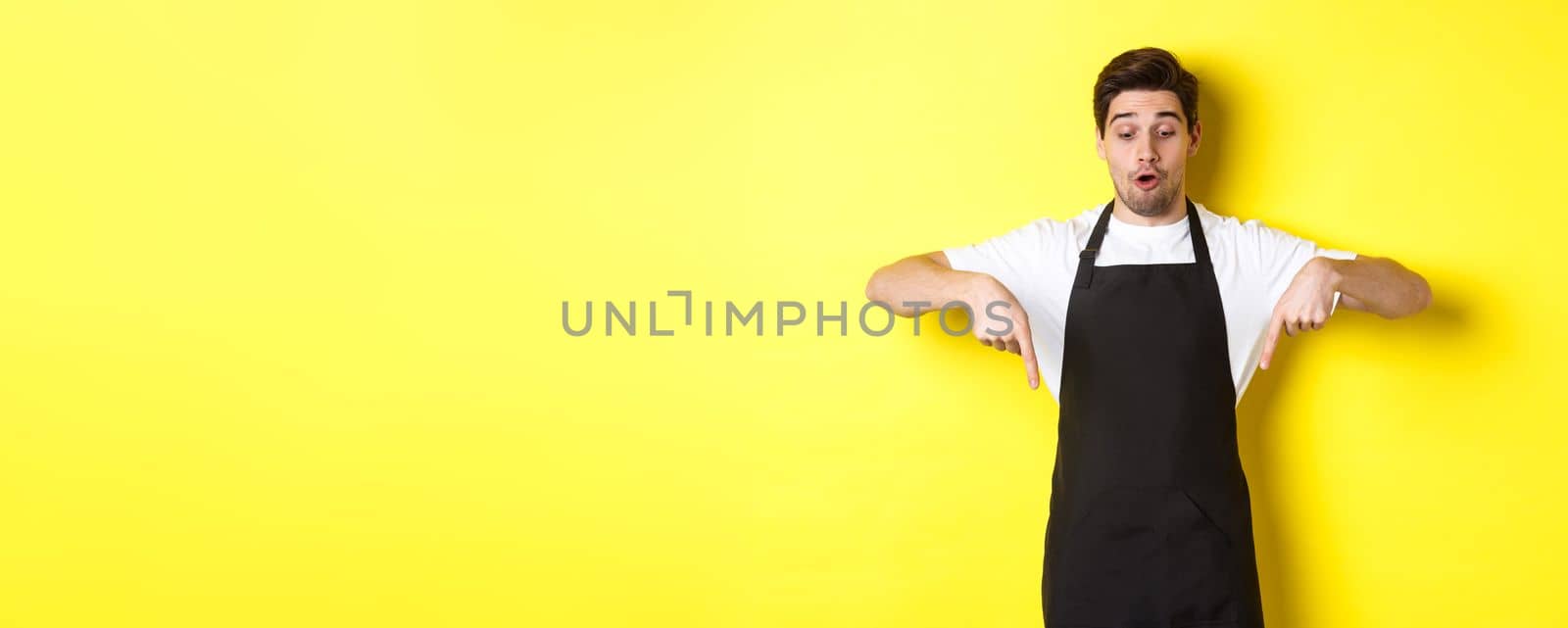 Surprised male barista in black apron checking out promo offer, pointing fingers down at logo banner, standing over yellow background by Benzoix