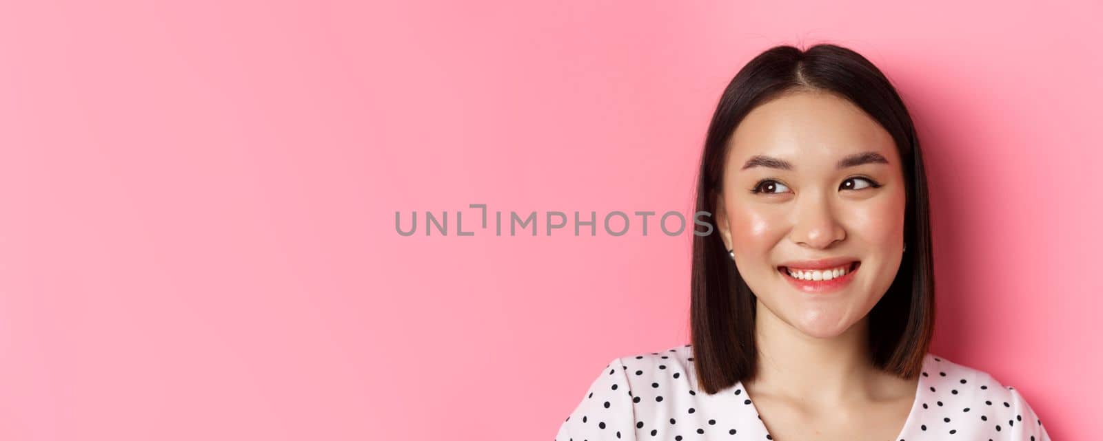Beauty and lifestyle concept. Headshot of pretty asian woman dreamy gazing left at copy space, smiling happy, standing over pink background by Benzoix
