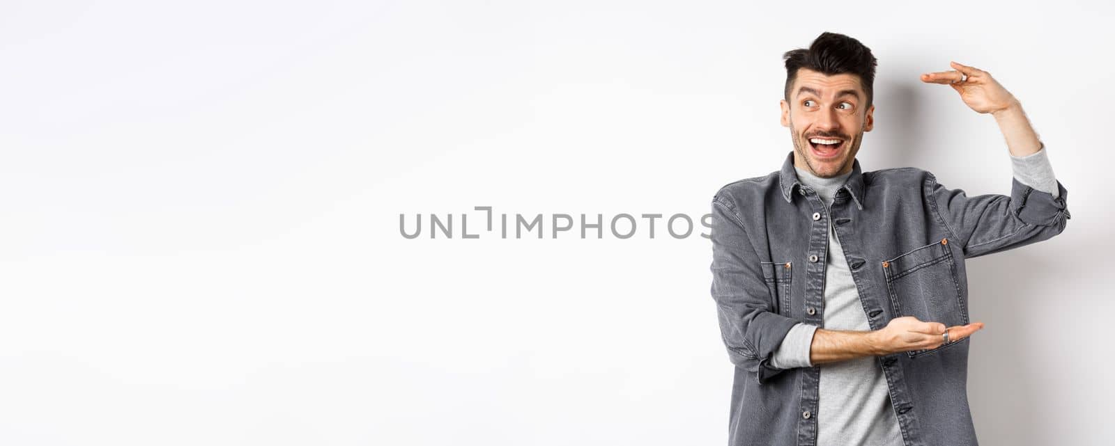 Excited guy looking at logo and showing big size thing, shaping large object and smiling amazed, standing in denim jacket on white background by Benzoix